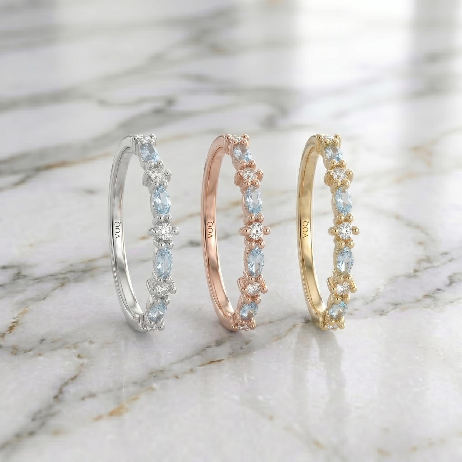 Three solid gold rings with aquamarine marquise and round moissanite stones are displayed on a marble surface.