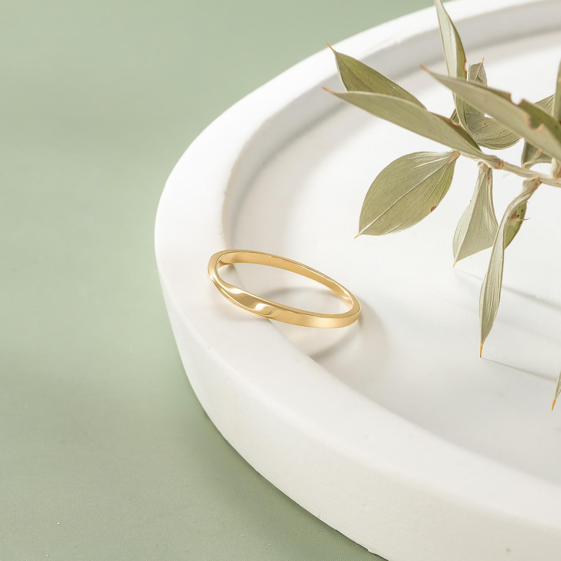 A gold ring rests on a white plate, accompanied by a branch of green leaves.