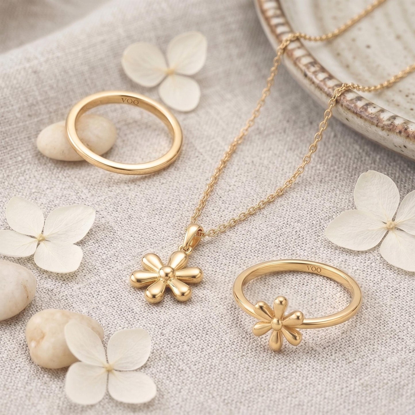 a collection of gold jewelry, including rings, a necklace, and a pendant, arranged on a beige fabric surface with white flower petals scattered around.
