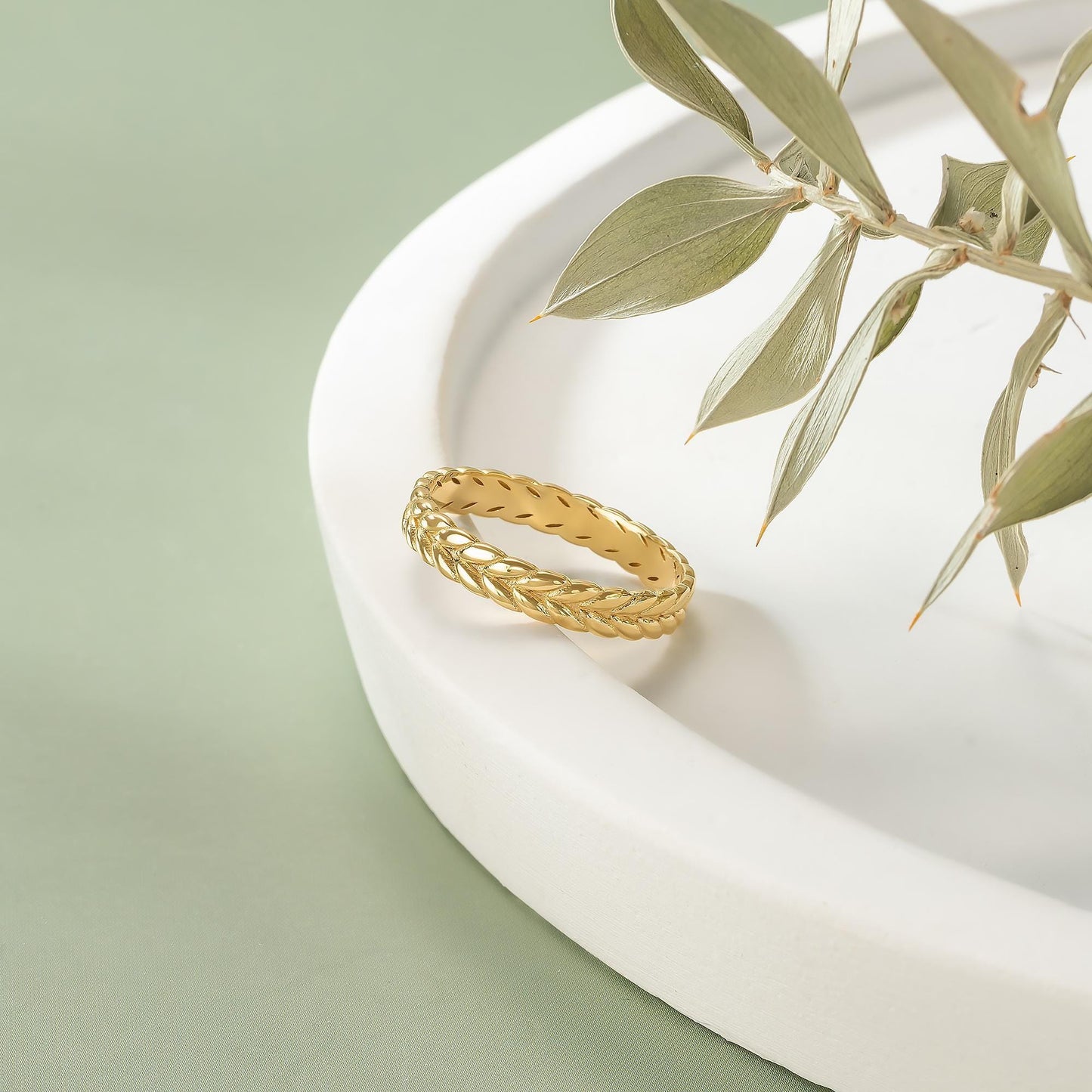 A gold ring with a braided design rests on a white plate, accompanied by a branch of green leaves.