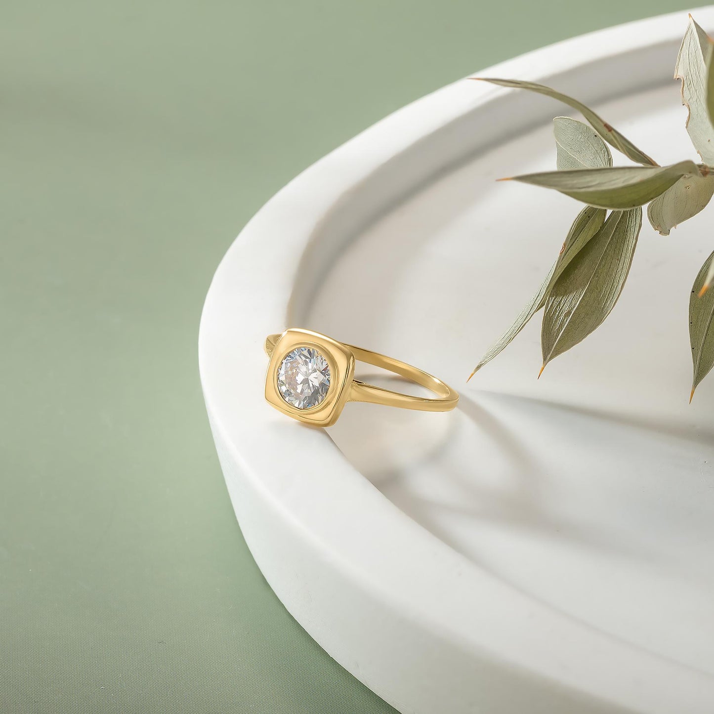 A gold ring with a large diamond sits on a white plate, accompanied by a green leaf.