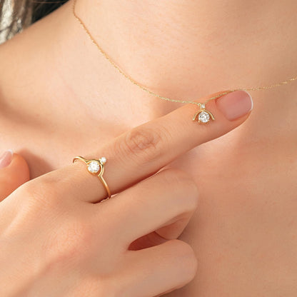 A close-up of a person's hand wearing a gold ring and necklace, with a small diamond pendant on the ring finger.
