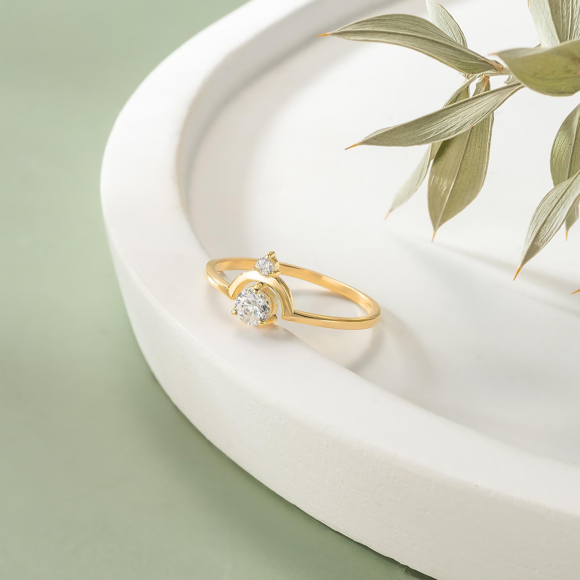 A gold ring with a large diamond sits on a white plate, accompanied by a plant with green leaves.
