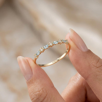 A gold ring with aquamarine marquise and round moissanite stones is being held in a person's hand.